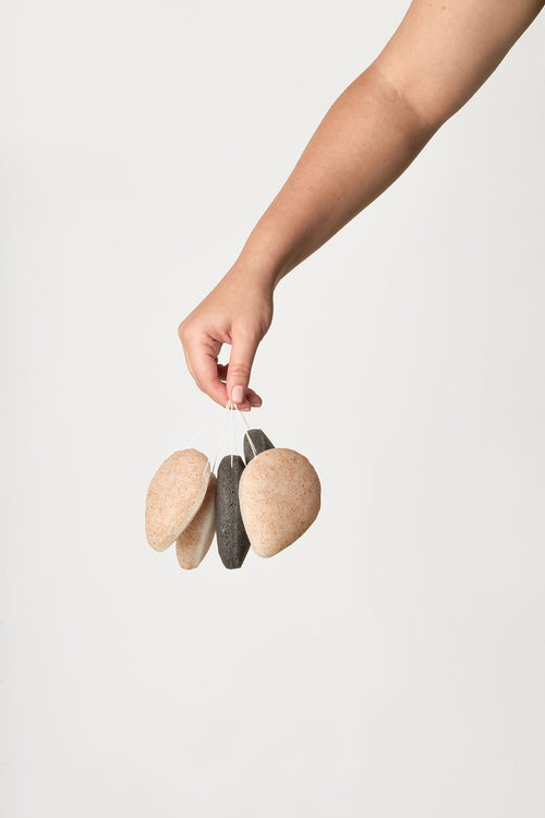 Hand holding Walnut and Charcoal Konjac Sponges by their strings against a neutral background
